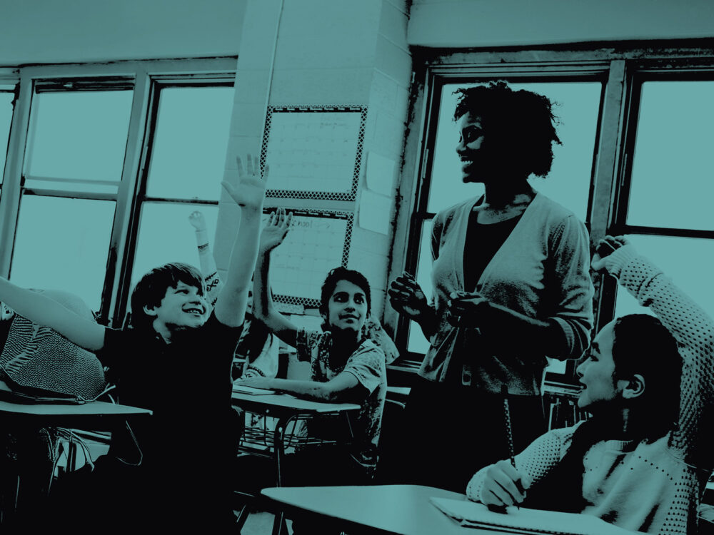 A female teacher leads a class of school children, some of whom have their hands raised. The image has a teal tint.