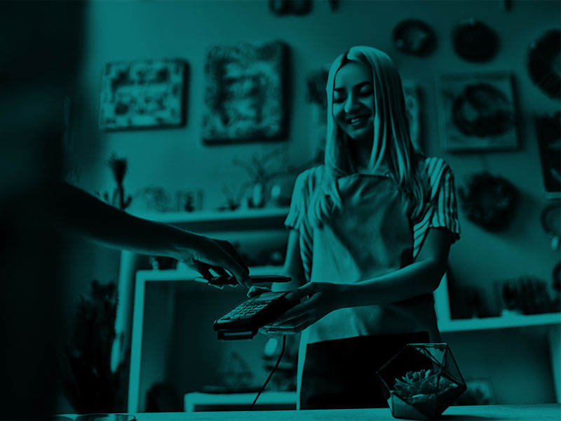 A woman holds up a card machine, while someone goes to tap their phone on said card machine. The woman is smiling. There is a green/blue filter over the photograph.