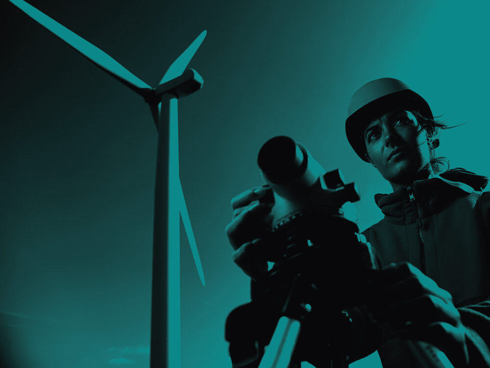 A worker wearing a hard hat uses equipment in front of a wind turbine. Teal filter.