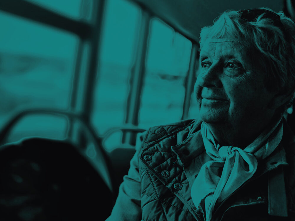An elderly woman looks out of a bus window. Teal tint.
