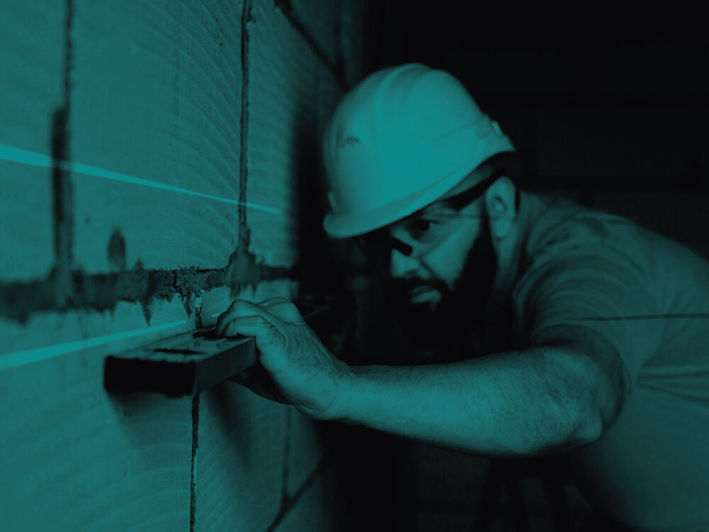 A builder uses a level against a wall. Image has a cyan tint.