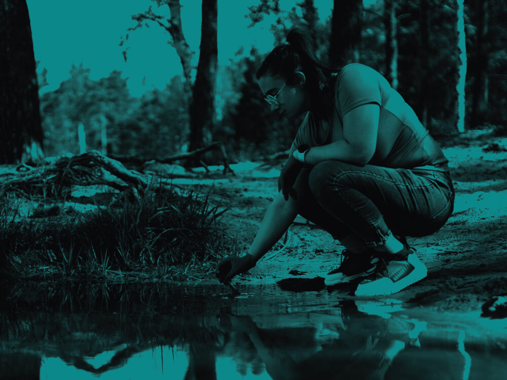 A woman crouches down taking a sample from a small body of water. The image has a teal tint.