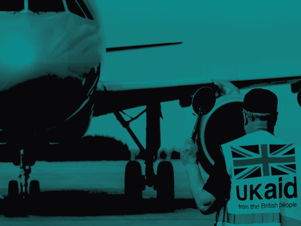 A man wearing a high-vis UKaid jacket guides a plane. Image has a teal tint.
