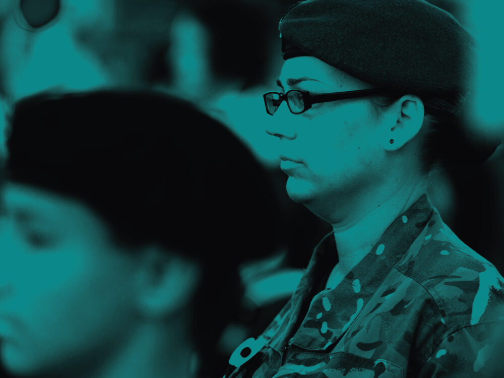 A soldier wearing glasses stands to attention, looking off the left of the screen. The image has a teal tint.